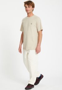 Beige katoenen t-shirt met korte mouwen en een ronde hals, voorzien van een klein zwart logo. Gepaard met crèmekleurige broek en donkere schoenen.