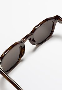 Braune Schildpatt-Sonnenbrille mit dunklen Gläsern, kantigem Rahmendesign und silberfarbenen Metallakzenten an den Schläfen.