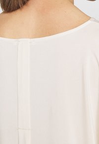Top in cotone beige chiaro con scollatura ampia, caratterizzato da una cucitura verticale sul retro e una consistenza morbida. Nessun motivo aggiuntivo o decorazioni.