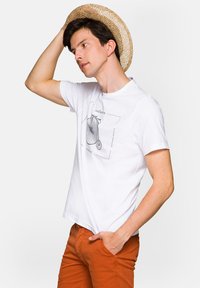 Biała bawełniana koszulka z grafiką roweru, wzbogacona czarnym napisem. Model nosi słomkowy kapelusz i rdzawobrązowe spodnie.