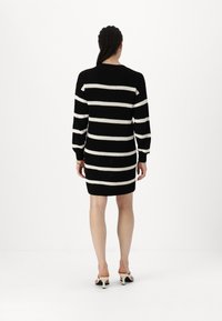 Vestido de malha riscado em preto e branco, com mangas longas, textura canelada e um ajuste justo, terminando acima da metade da coxa, usado com sapatos de salto aberto.