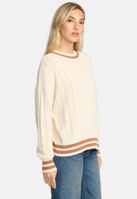 Pull crème avec des côtes verticales, présentant des accents rayés marron au niveau de l'encolure et de l'ourlet. Fabriqué dans un tissu doux ettexturé.
