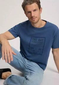 Niebieska bawełniana koszulka z krótkim rękawem z przodu ma graficzny nadruk w ciemnym kolorze, zestawiona z jasnoniebieskimi dżinsami.