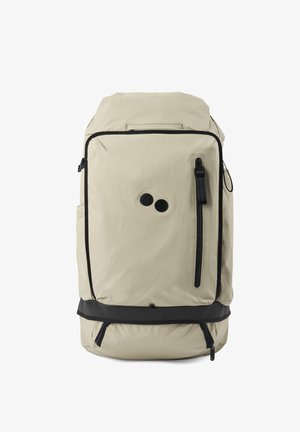 Beiger Stoff-Rucksack mit schwarzen Akzenten, ausgestattet mit einer vorderen Reißverschlusstasche, runder Form und doppeltem kreisförmigem Emblem auf der Vorderseite.