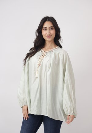 Vrouw met lang donker haar, gekleed in een losse, lichtgroene blouse met borduurwerk en franjes, gecombineerd met donkerblauwe jeans, staand tegen een effen achtergrond.
