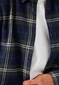 Marinblå och grön rutig flanellskjorta med blå knappar, lager på lager över en vit t-shirt med rund hals. Har en mjuk textur och en normal passform.