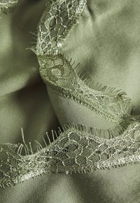Tissu satiné vert olive clair avec bordure en dentelle délicate présentant des motifs floraux et des détails de fils fins, drapé en plis doux.