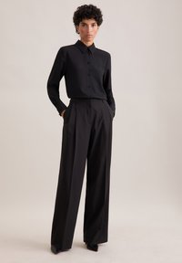 Camicia nera con bottoni e colletto abbinata a pantaloni neri a gamba larga. Il tessuto appare liscio, con dettagli sartoriali e tasche laterali.