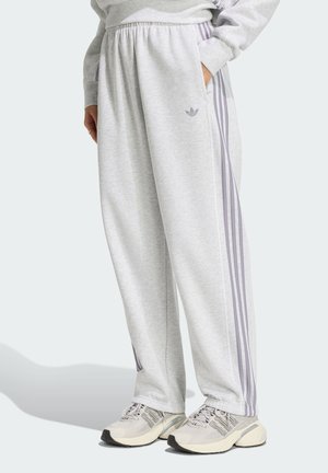 Osoba ubrana w jasnoszare spodnie dresowe Adidas z fioletowymi paskami i logo, w połączeniu z kremowymi butami sportowymi, stojąca z jedną ręką w kieszeni.
