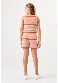 Kind dat met de rug naar de camera staat, in een bijpassende gestreepte mouwloze top en shorts, met witte sneakers, tegen een witte achtergrond.
