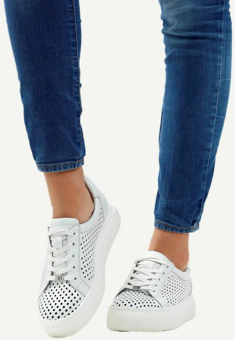 Personne portant un jean bleu et des baskets en cuir blanc perforé avec des semelles épaisses, debout devant un fond uni.