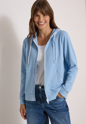 Femme souriante portant un sweat à capuche zippé bleu clair sur une chemise blanche et un jean bleu, debout avec une main dans la poche devant un fond uni.