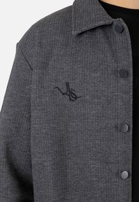 Chaqueta gris texturizada con botones, que cuenta con cuello, botones negros y un sutil bordado de logo negro en el pecho.