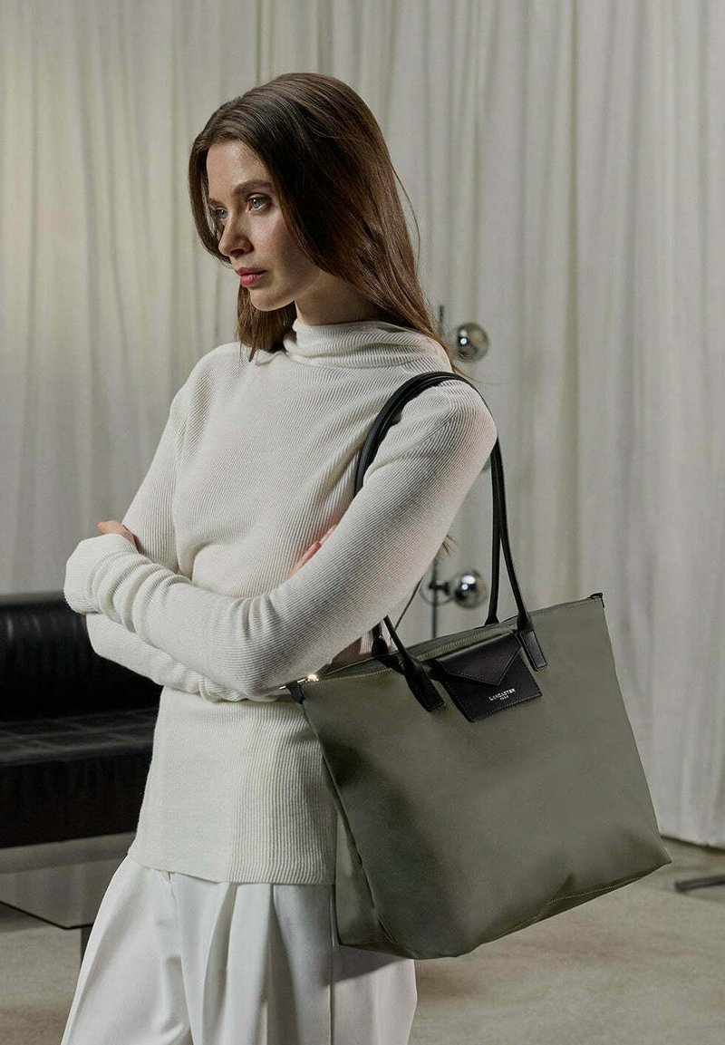 Borsa tote color khaki con dettagli in pelle nera, caratterizzata da una forma strutturata e due manici lunghi. La borsa ha una trama liscia e un design minimale.