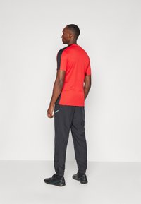 Rotes Kurzarmhemd mit schwarzen Akzenten, kombiniert mit schwarzen Jogginghosen mit einer Reißverschlusstasche und elastischen Bündchen, getragen mit schwarzen Sportschuhen.