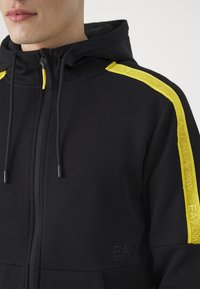 Detailní záběr na černou mikinu na zip s kapucí, černými šňůrkami a žlutými pruhy se značkou na ramenech s nápisem "EA7 Emporio Armani."