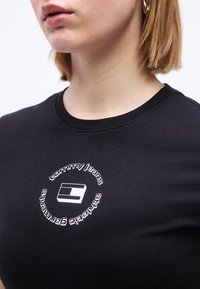 Γυναίκα που φοράει μαύρο μπλουζάκι με λευκό κυκλικό λογότυπο "tommy jeans authentic garments" στο στήθος και χρυσό κρίκο στο αυτί, ανοιχτόχρωμο δέρμα, κοντά μαλλιά.