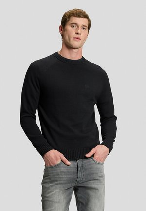 Mann steht vor schlichtem Hintergrund, trägt einen schwarzen Pullover und hellgraue Jeans mit den Händen in den Taschen, schaut nach vorne.
