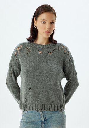 Grijze gebreide trui met versleten gaten, ronde halslijn en geribde zoom. Draagt over lichtblauwe denim voor contrast.