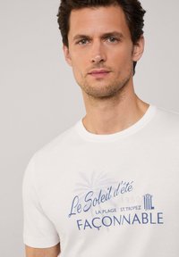 Camiseta de algodón blanca con cuello redondo, mangas cortas y texto gráfico azul con motivos de palmeras y arquitectónicos.