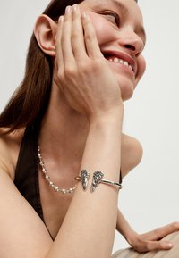 Pulsera de plata con detalles en forma de ala, de forma circular y superficie texturizada. Se lleva en la muñeca junto a un collar de cuentas de plata.