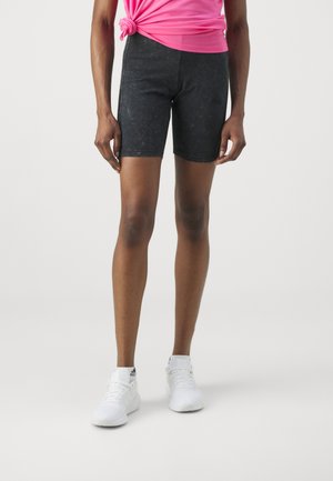Personne portant un short de sport noir, des baskets blanches et un t-shirt rose noué à la taille, sur un fond uni.