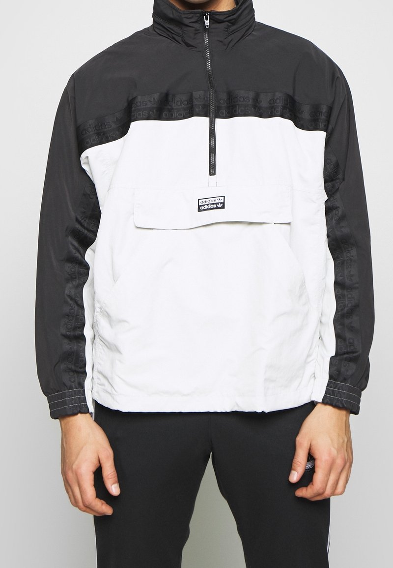 White and black zip-up pullover jacket, with a prominent front pocket, logo patch, and textured black sleeves featuring repeating brand name.
