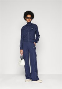 Tuta navy realizzato in morbido tessuto con strisce bianche sui lati. Include una giacca con zip e pantaloni larghi, abbinati a una piccola borsa bianca.