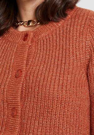 Primer plano de una mujer que lleva un cárdigan de punto texturizado color óxido con grandes botones y un collar grueso de cadena dorada.