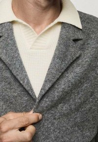 Szary wełniany płaszcz z klapami, nałożony na teksturowany biały sweter. Ręka trzymająca przód płaszcza, ukazująca miękkość tkaniny.