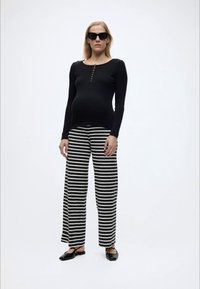 Έγκυος γυναίκα που φοράει μαύρο μπλουζάκι με μακριά μανίκια, μαύρο και άσπρο ριγέ παντελόνι με φαρδιά πόδια, μαύρες μπαλαρίνες και σκούρα γυαλιά ηλίου σε λευκό φόντο.