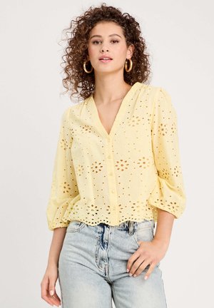 Frau mit lockigem Haar trägt gelbe Bluse mit Lochstickerei, durchgehenden Knöpfen und dreiviertellangen Ärmeln sowie hellblaue Jeans mit hoher Taille.