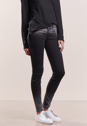 Vaqueros ajustados en negro degradado combinados con zapatillas blancas y una camiseta negra de manga larga, posando contra un fondo claro y liso.