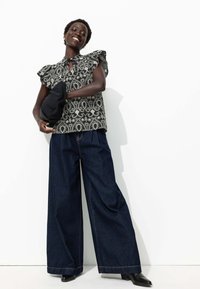 Blouse paisley noir et blanc avec des manches volantées, associée à un pantalon en denim sombre à jambes larges et des bottines noires. Fond blanc uni.