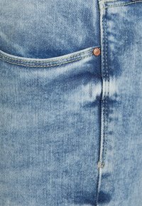 Vaqueros de mezclilla azul claro con un bolsillo frontal que presenta costuras en contraste y herrajes de color cobre, mostrando una textura desvaída.