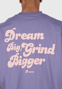 Lilla t-shirt med stort, kursiv hvid tekst på ryggen, der står "Dream Big, Grind Bigger" med mærkenavnet "Dropsize" nederst.