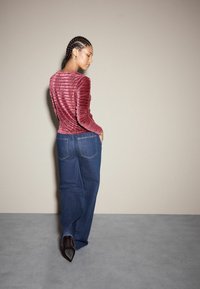 Frau mit geflochtenem Haar, die ein strukturiertes pinkfarbenes Langarmoberteil, weit geschnittene blaue Jeans und schwarze spitze Absätze trägt, und vor einer beigen Wand steht.