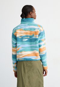 Pullover em fleece com um padrão de ondas em azul, laranja e creme. Apresenta um colar alto e mangas longas, com um corte descontraído.