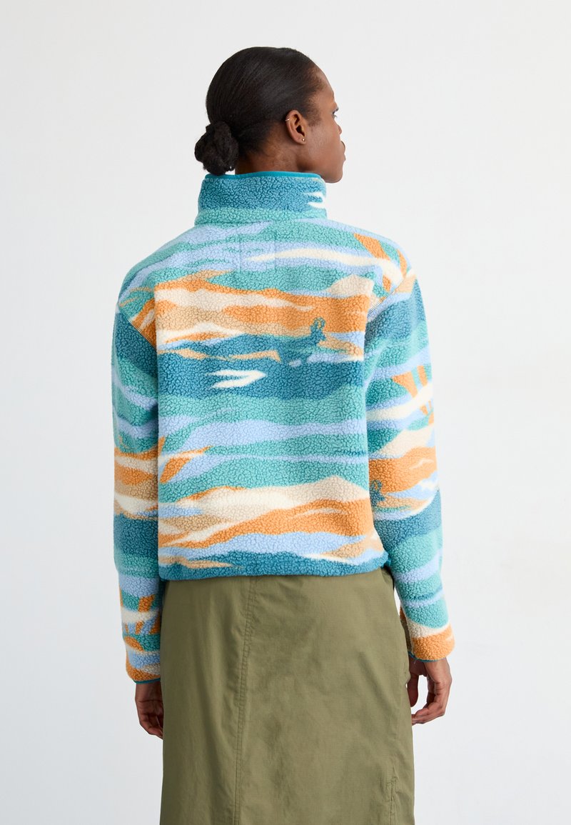 Pull en polaire bleu, orange et crème avec un motif de vagues. Il présente un col montant et des manches longues, avec une coupe décontractée.