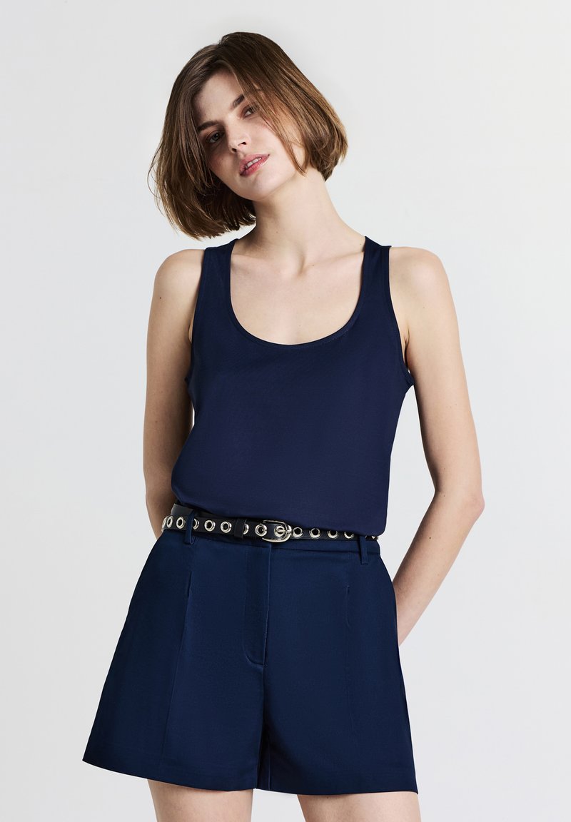 Canottiera navy realizzata in tessuto liscio, abbinata a pantaloni corti navy sartoriali con una cintura nera dotata di dettagli in grommet argentati.