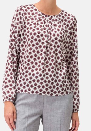 Blouse blanche à manches longues avec motif géométrique rouge et violet, associée à un pantalon gris clair sur mesure.