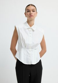 Witte zonder mouwen blouse met een kraag, sluiting met knopen aan de voorkant, getailleerde pasvorm en licht uitlopende zoom. Draagt met zwarte, op maat gemaakte broek.