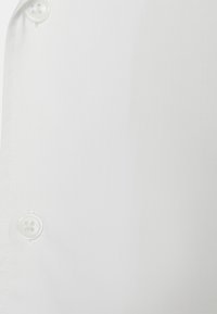 Bílá látka košile s hladkým povrchem. Tři bílé knoflíky viditelné u okraje, s čistým a ostrým vzhledem.