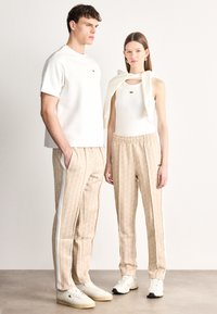 Λευκό πουκάμισο με κοντά μανίκια, κρεμ τοπ, μπεζ παντελόνι με σχέδια και λευκές ρίγες, και λευκά αθλητικά παπούτσια με υφασματικές λεπτομέρειες.