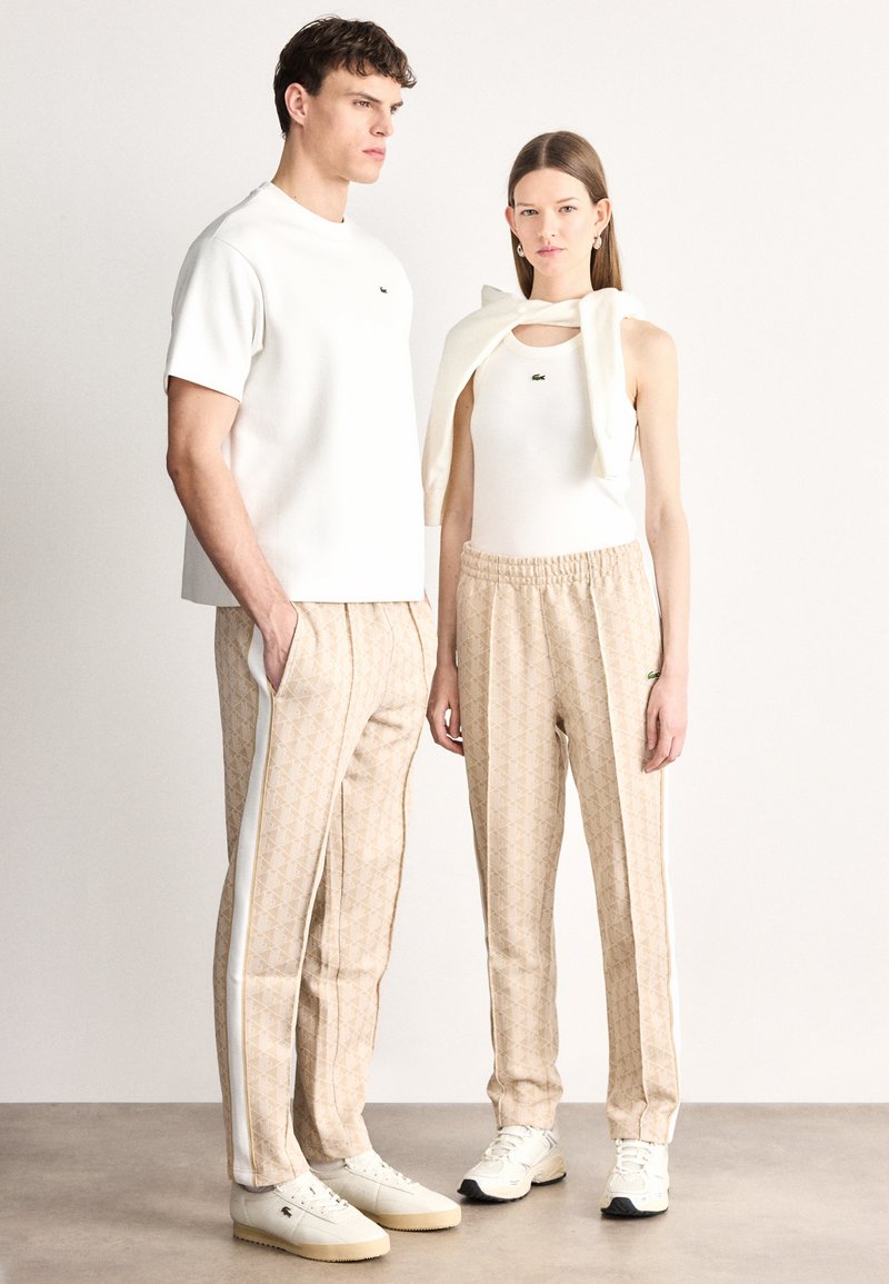 Camicia bianca a maniche corte, canottiera color crema, pantaloni beige con motivo a strisce bianche e sneakers bianche con dettagli strutturati.