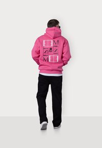 Sweatshirt à capuche rose avec un texte graphique noir et un motif floral dans le dos, porté avec un pantalon noir et des baskets blanches.
