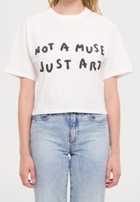 Vit kortärmad t-shirt med svart text "ÄR INTE EN MUS, BARA KONST" bärs tillsammans med ljusblå högmidjade jeans på en person.