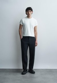 Νέος άνδρας με σγουρά σκούρα μαλλιά στέκεται μπροστά σε απλό φόντο, φοράει ένα λευκό κοντομάνικο πουκάμισο, μαύρο παντελόνι και μαύρα παπούτσια.