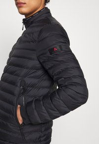 En person som bär en svart vadderad jacka med dragkedjefickor och en logolapp på vänster överarm, med handen i höger ficka.