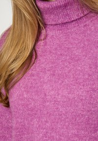 Adīta augsto apkakli džemperis no mīksta, viegla rozā auduma ar strukturētu virsmu. Iezīmējas ar augstu, ribotu apkakli.